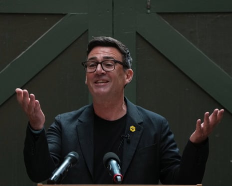 Andy Burnham in glasses and a dark suit gestures with both hands raised while speaking at microphones