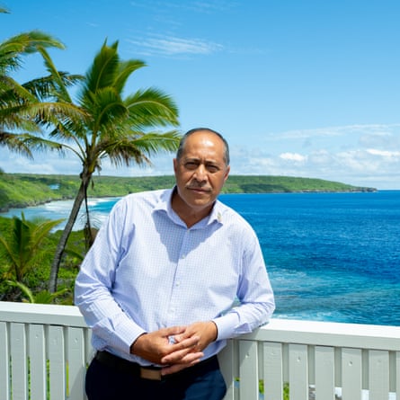 A man leans on a white fence in front of a tropical backdrop with palm trees and blue sea