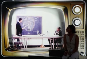 A woman sits in front of a projection of a 1950s-60s television set showing a programme from the period