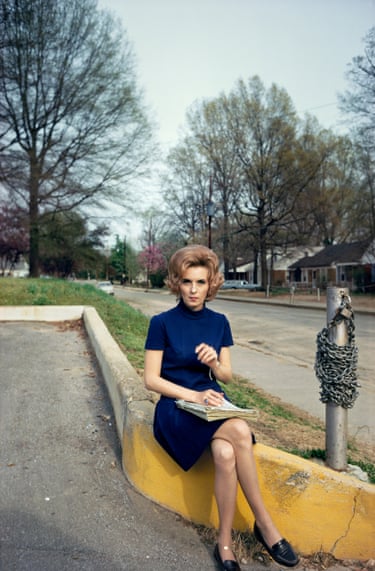 A woman with light brown hair wearing a dark blue polo neck dress sitting on a wall next to a silver pole with a chain around it. Trees are in the background