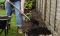 A person seen from the waist down adding compost to a garden bed using a spade and wheelbarrow