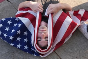 A demonstrator lies on their back on the ground with the US flag draped around their head and upper body