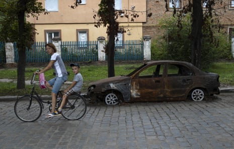 A mother and child ride a bike past a destroyed car in Orikhiv, Zaporizhzhia oblast