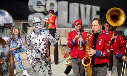 Performers at the opening event: a brass band including saxophone, trumpet, trombone and french horn play, the musicians wearing red military-style jackets, while mime artists in silver costumes entertain people as they enter the arena