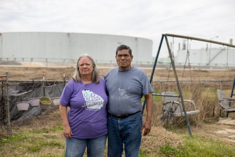 A middle-aged man and woman stand near a swing set with an industrial facility in the background.