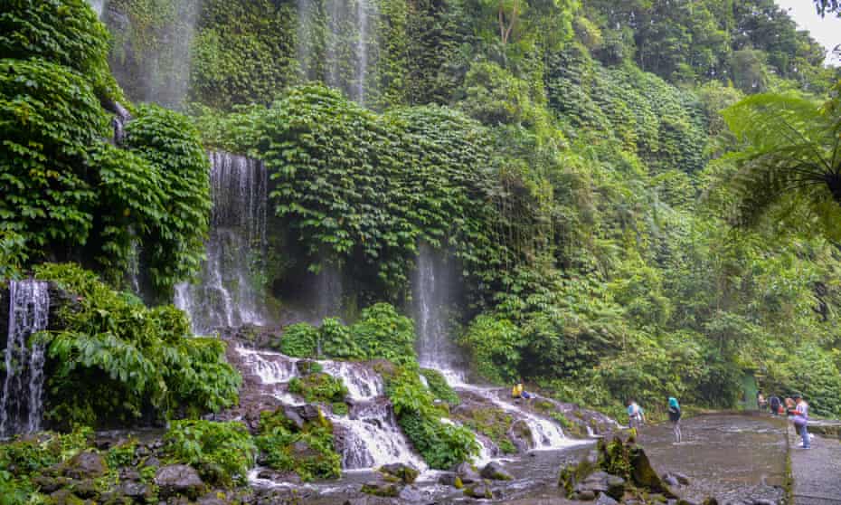 Tourists stand in front of a waterfall set into a lush green hillside.