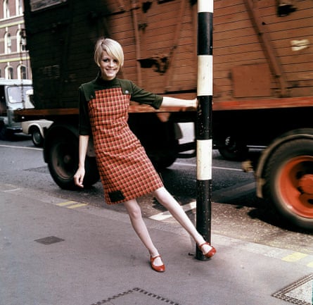 Twiggy in 1966, smiling, wearing a red checked dress, hanging off a lamp-post as a lorry drives past