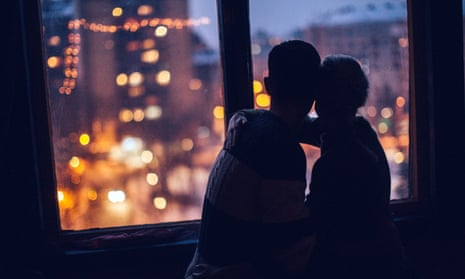 A silhouette of a couple embracing at dusk as they looking out a window on to a cityscape
