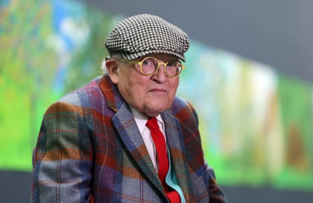 An older man wearing a tartan blazer, red tie, black and white gingham hat and round, yellow glasses in front of a painting.