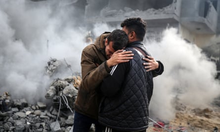 Aftermath of an Israeli airstrike in Gaza, with a man comforting another man in front of a smoking ruin