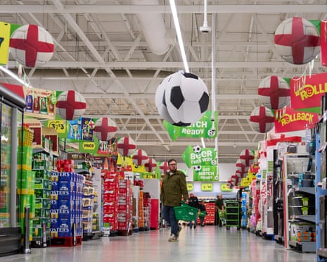 A man holds a green shopping basket as he walks down a supermarket aisle displaying football-related promotions.