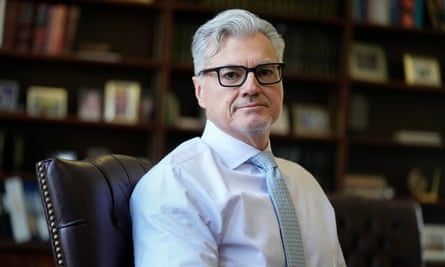 Middle-aged Latino man with thick white hair and black-framed glasses, in shirt and tie, sits in leather chair with extensive bookcases behind him.