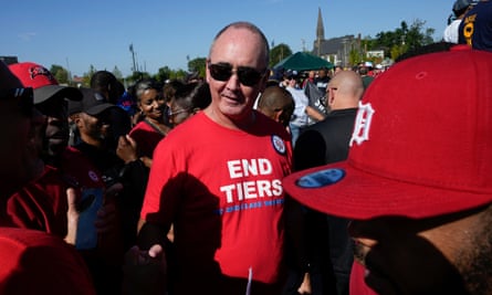 In the middle of a crowd of people who are mostly Black, a middle-aged white man with mostly no hair wears sunglasses and looks into the sun, wearing a red T-shirt that says ‘End Tiers’.