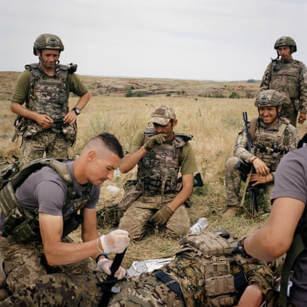 A group of soldiers sit in a field performing a drill using medical equipment on what appears to be a colleague lying on the grass