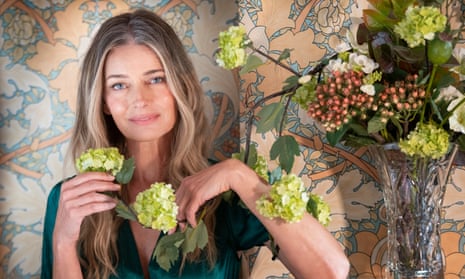 Model, actress and author, Paulina Porizkova, with floral wallpaper behind her and a vase of flowers to one side. She's holding two hydrangea blooms.