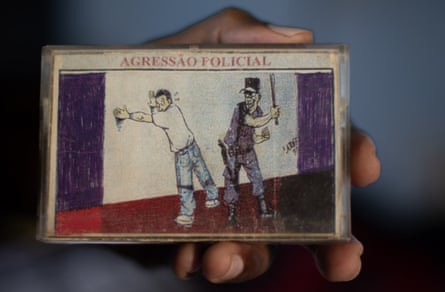 A hand holds a small card with a drawing of a police officer striking a man, titled “AGRESSÃO POLICIAL” (police aggression), with simple coloured blocks in the background.