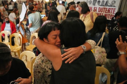 Two women hug in a hall filled with people holding signs calling for Duterte to face justice