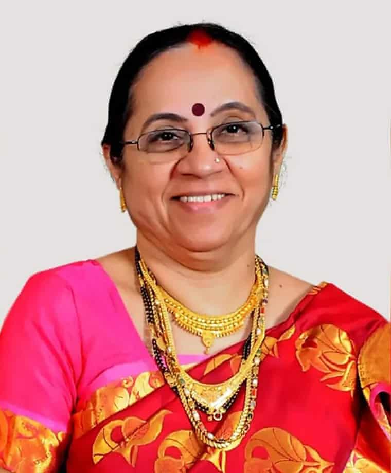 A smiling older Indian woman, wearing glasses and a red sari, with layered gold necklaces