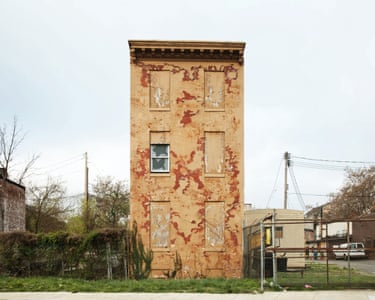 a tall thin house, mottled ochre and red, most windows boarded, on an urban street corner