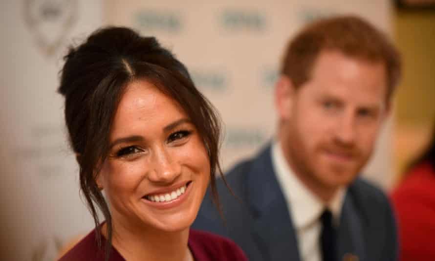 Closeup of Meghan smiling at someone off camera with Prince Harry out of focus in the background