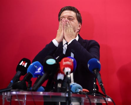 Benoît Payan standing at lectern behind press microphones puts both hand to his mouth in preparation to blow a kiss