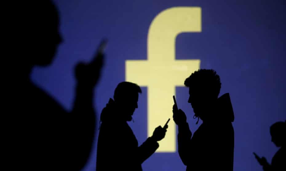 Silhouettes of mobile users are seen next to a screen projection of Facebook logo.