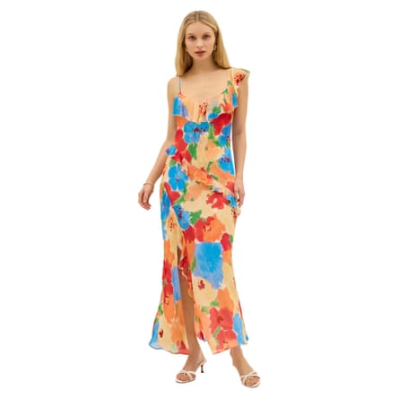 A model wears a maxidress that has a floral watercolour pattern. The pattern is red orange bright blue and peach tones. There is a frill neckline. The right shoulder has a thin strap and the left a cap sleeve. The model wears the dress with white heeled spaghetti strapped shoes.
