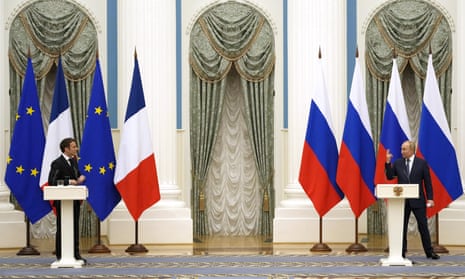 Russian President Vladimir Putin, right, gestures at French President Emmanuel Macron as they stand at well-spaced white lecterns. Behind Putin are four Russian flags. Behind Macron are two French flags and two European Union flags.