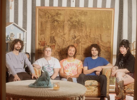 Wednesday sit on a faded sofa and chairs with a faded tapestry-type hanging in the background and table in foreground.