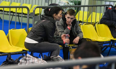 A woman and a boy sit on yellow seats surrounded by bags in a sports hall in Budapest.