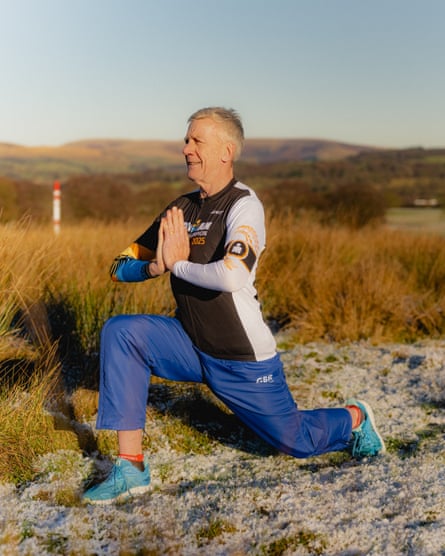 A man wearing dark-blue tracksuit bottoms, a sporty brown top and blue trainers, performing a yoga lunge on some shrubland
