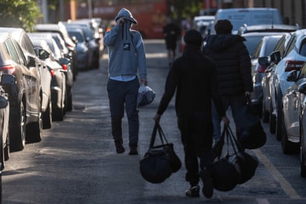 Three people carry bags as they walk along the road between parked cars