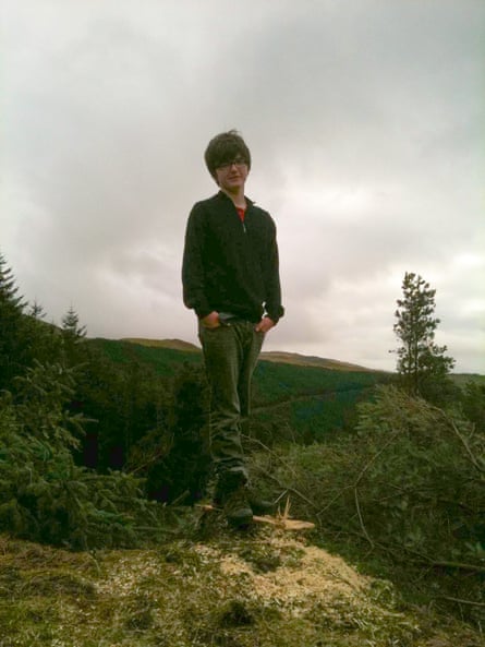 He stands on a tree stump in a woodland setting with hills behind him