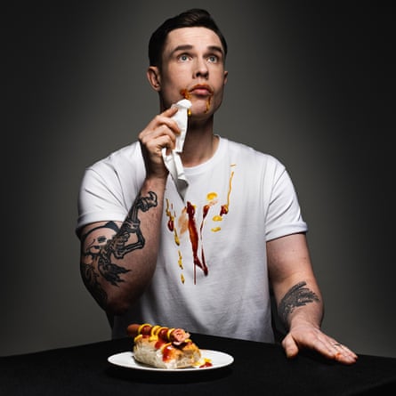 Comedian Ed Gamble wipes sauce from his mouth, his T-shirt stained, a hot dog on the plate before him