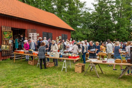 People buying things displayed off trestle tables outside a large hut in Sweden with trees behind