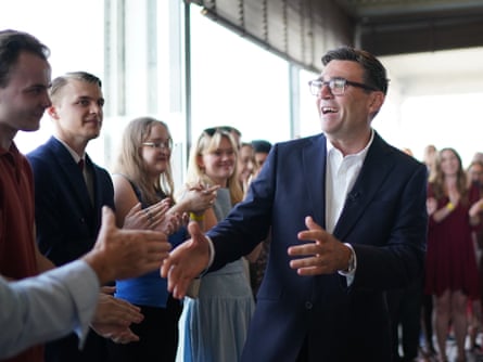 Andy Burnham smiles and greets a line of people standing in front of a window, with his hands held out.