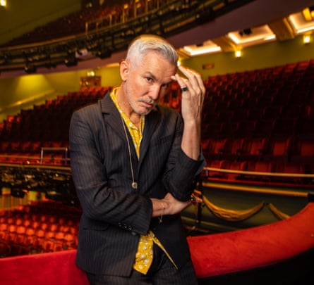 A man in a grey suit and a yellow shirt, standing in a theatre full of plush red velvet seats