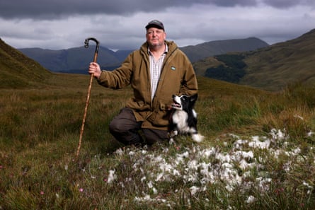 A man kneels next to a black and white dog, holding a shepherd's crook in one hand. In front of them fleece is scattered across the grass