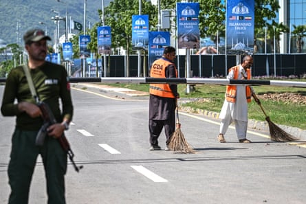 A man with an automatic rifle stands in a road as two others sweep with brushes