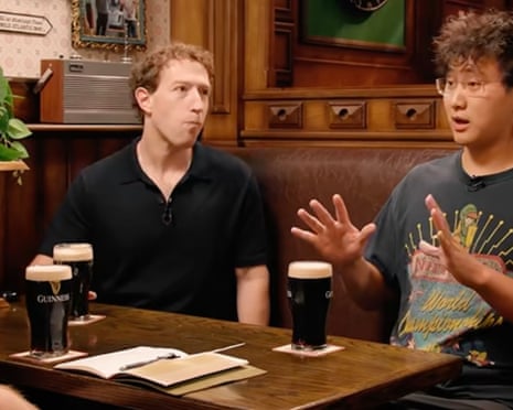 A man wearing a black short sleeve shirt pulls an uncomfortable face while holding a pint of Guinness on a table with two other full pints