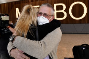 A man with glasses and a white mask hugs a woman with blonde hair inside Melbourne airport