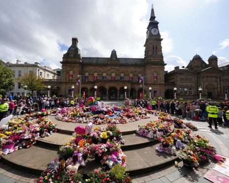 A large crowd gathers around a circular memorial covered in flowers, stuffed toys, and tributes in Southport