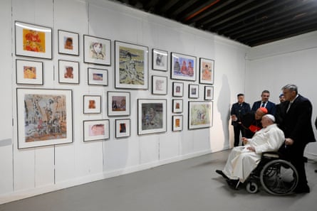 Pope Francis views a wall of small, framed paintings at the Giudecca’s women’s prison: he is seated in a wheelchair while attendants look on