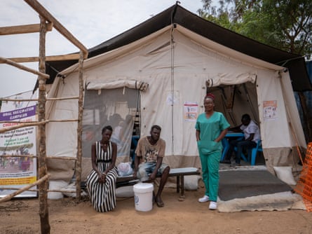 Three people, one in a nurses’ scrubs outside a tent