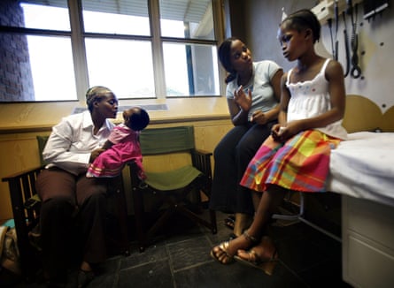 A young black African woman sits on a bed talking to a young black girl next to her as another woman plays with a baby