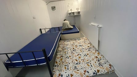 A man sits facing away from the camera on a bed in a small room almost filled with three beds