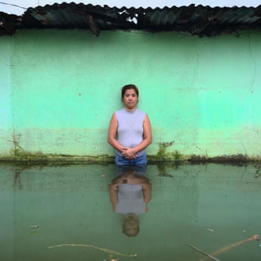 A woman stands by a green wall thigh-deep in flood water