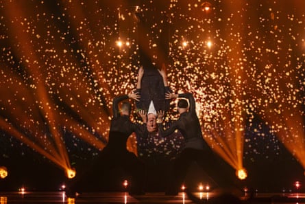 Man in black suit is lifted upside down on stage with lots of gold lights