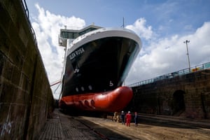 Greenock, Scotland. People in hard hats and red or blue overalls can be seen next to the front section of the hull of a large vessel in a dry dock