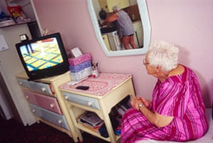 an older woman in a pink striped dress sits in a room with pastel-coloured walls and furniture watching television. a second figure is visible in a mirror on the wall